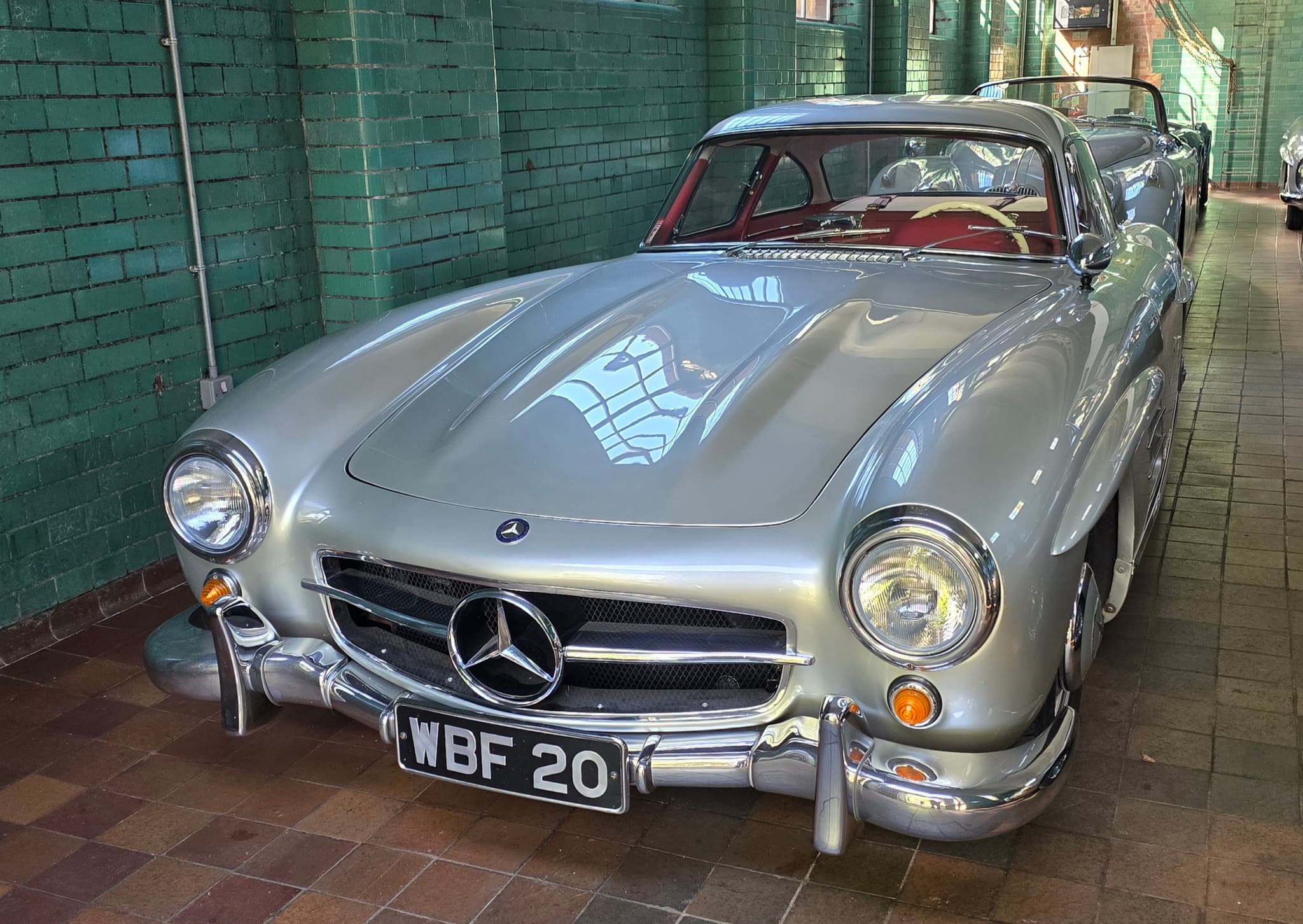 A silver Mercedes-Benz 300 SL coupé with round headlamps and a wide grille on a brick floor against green tiles.