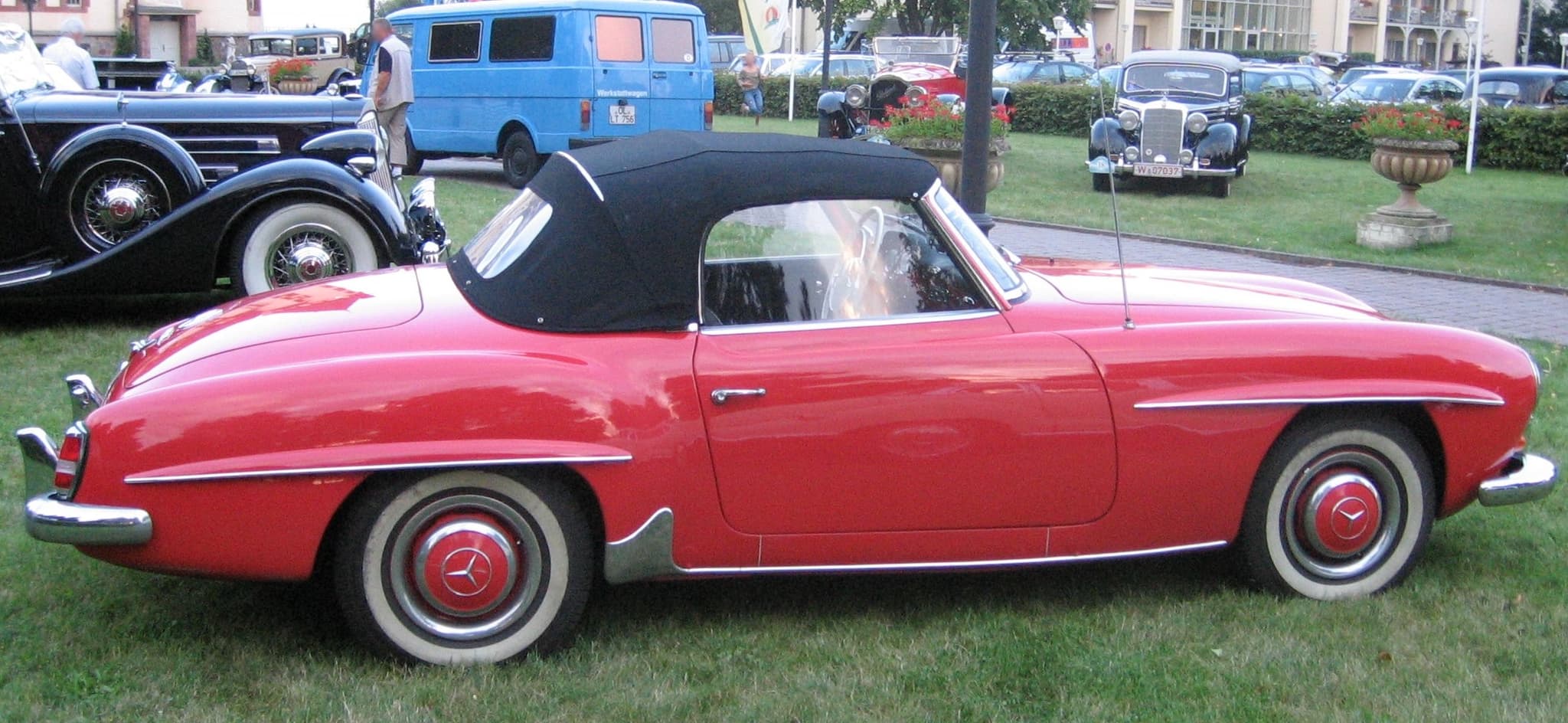 A red Mercedes-Benz 190 SL roadster with its black hood raised, seen from the side on grass at a car meeting.