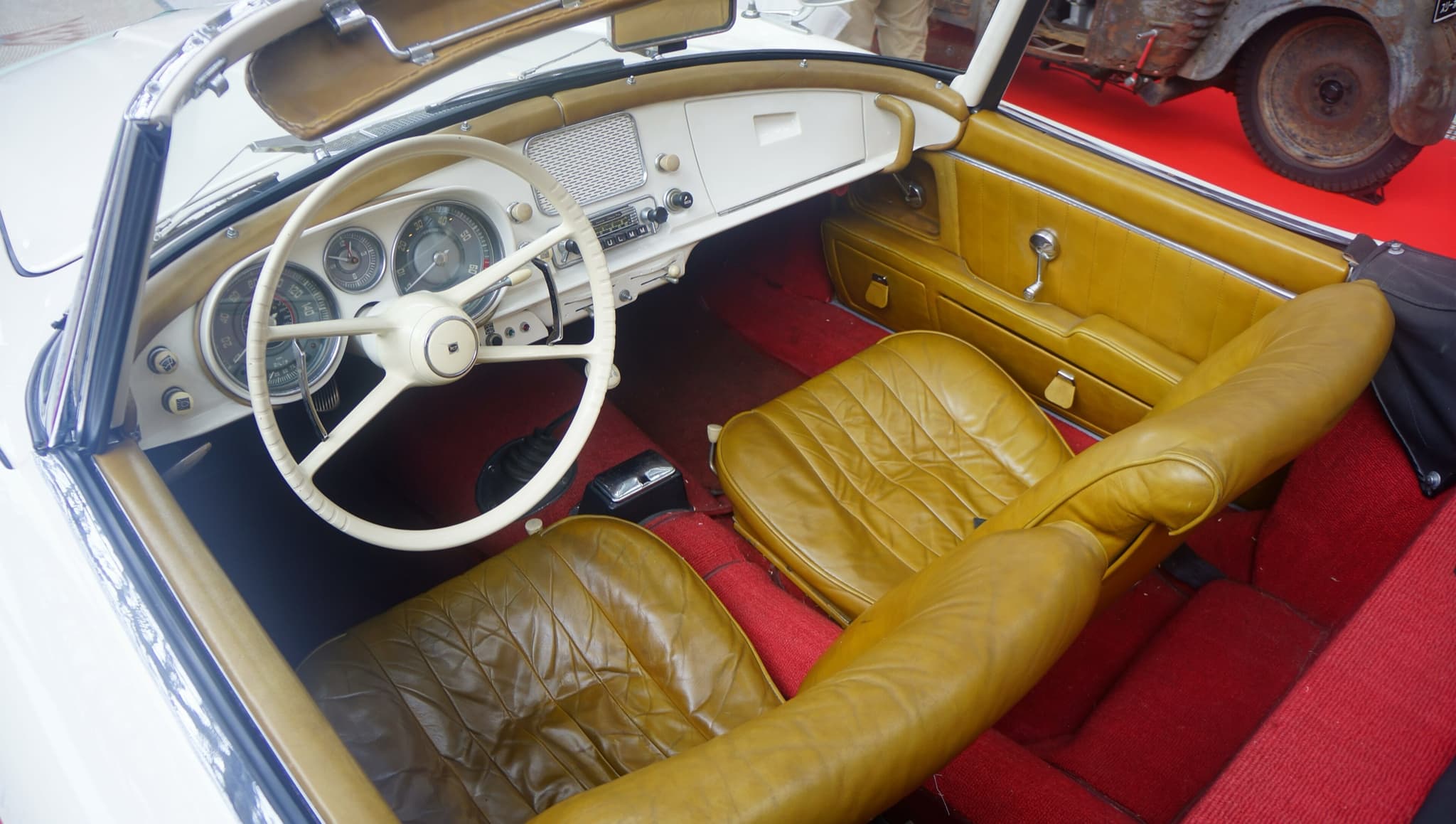 The cockpit of a white BMW 507 with mustard leather seats, an ivory steering wheel and a loudspeaker grille in the middle of the dashboard. Opens a larger view