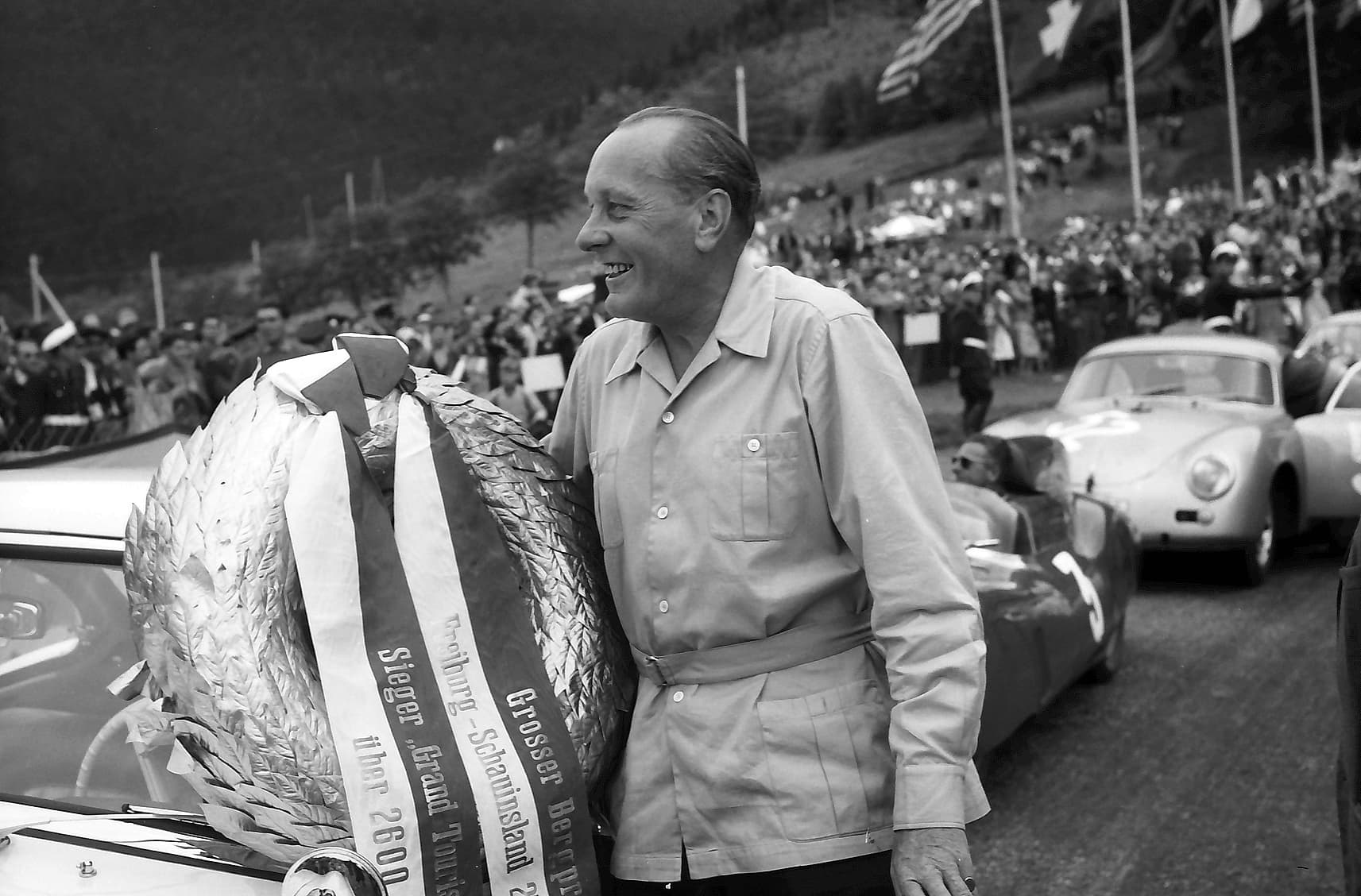 A black-and-white photograph of Hans Stuck smiling and holding a large laurel wreath with ribbons beside a car at a hill climb.