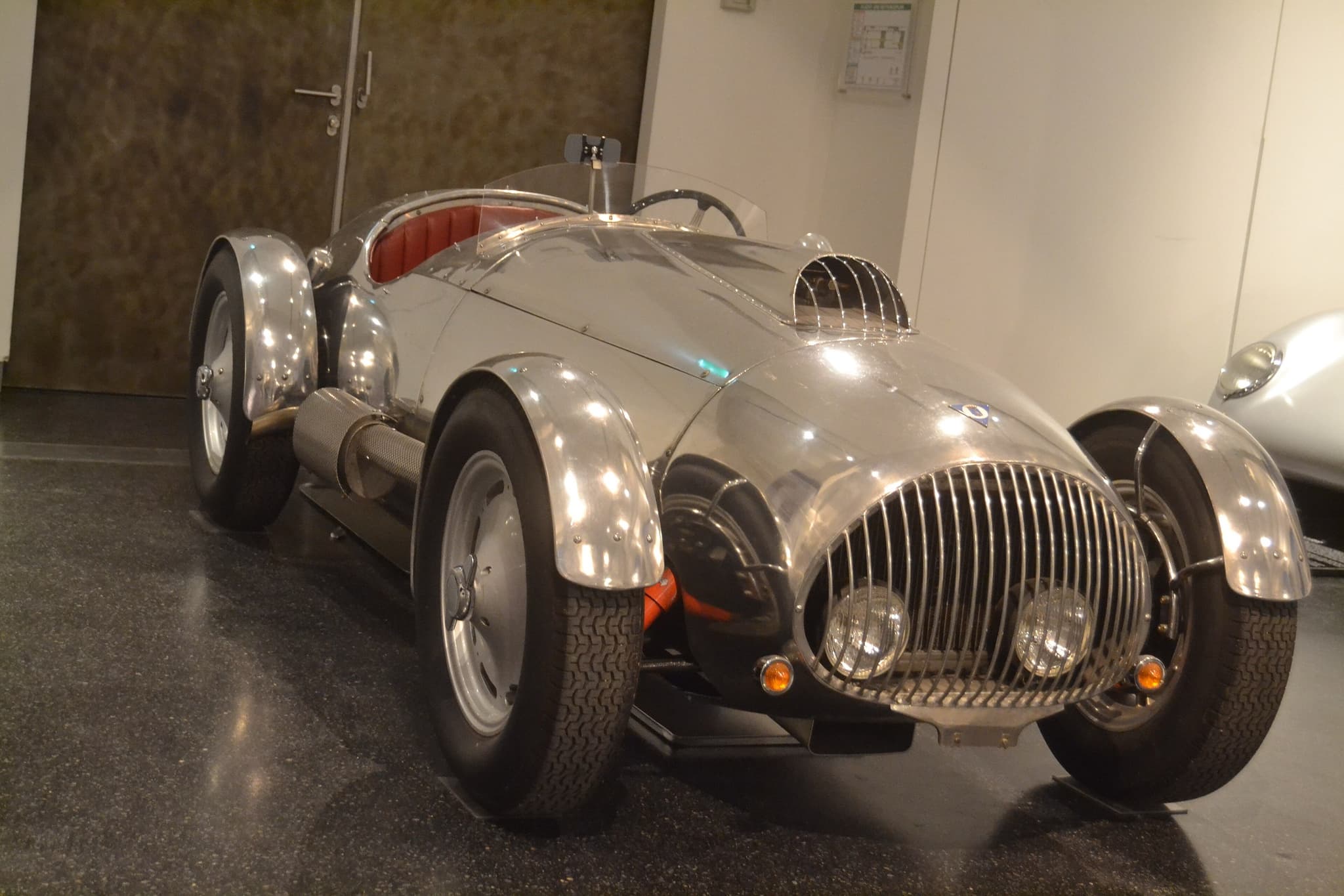 A polished aluminium AFM open racing car with a curved barred grille and cycle wings on a museum floor.