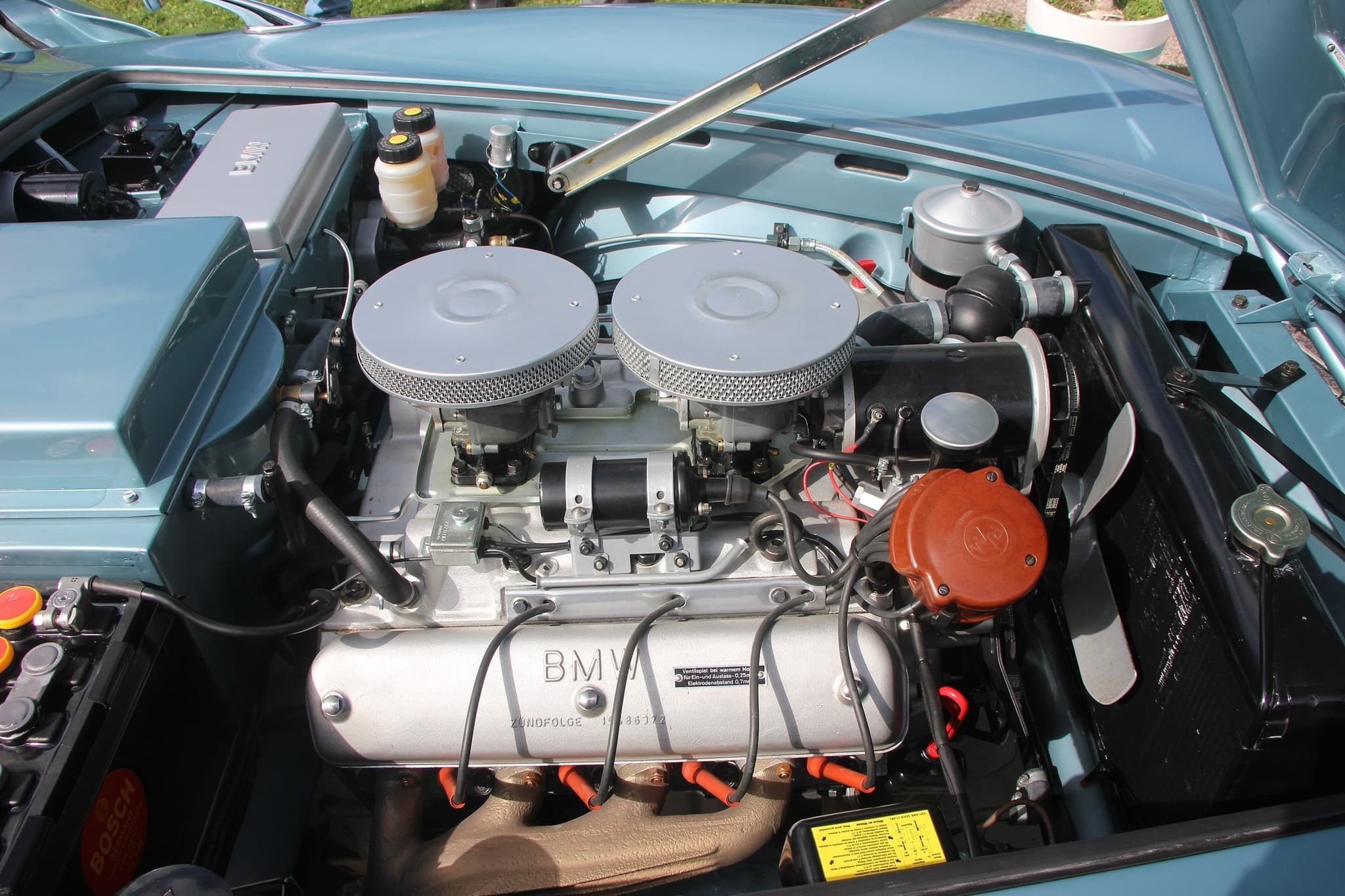 The engine bay of a pale blue BMW 507: an aluminium V8 with BMW rocker covers, two round air cleaners and black plug leads with orange boots. Opens a larger view