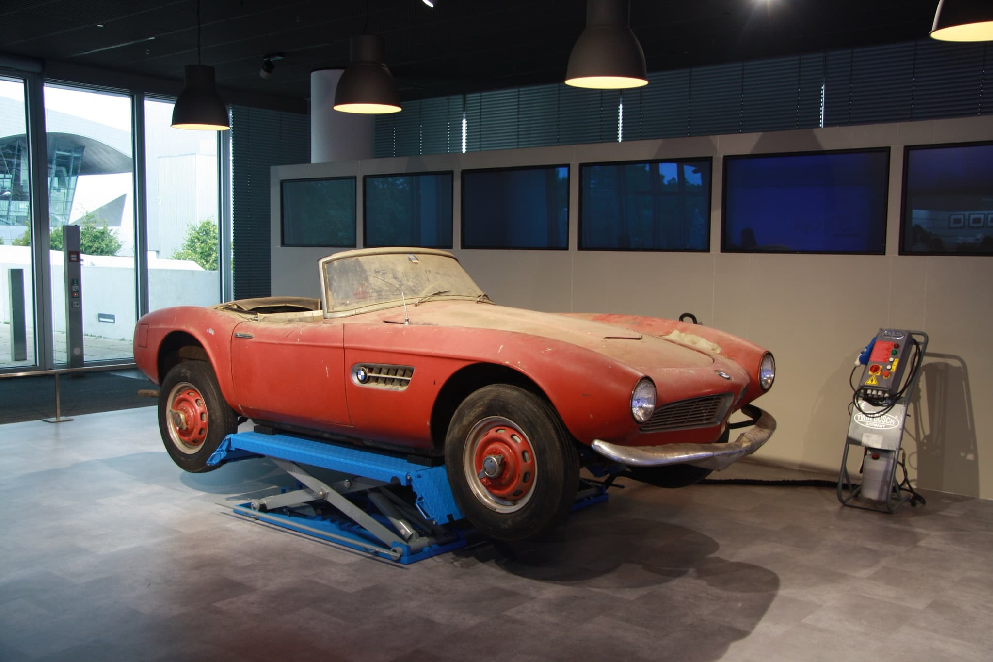 A dusty red BMW 507 with faded paint and red wheels on a blue lift in a museum hall. Opens a larger view