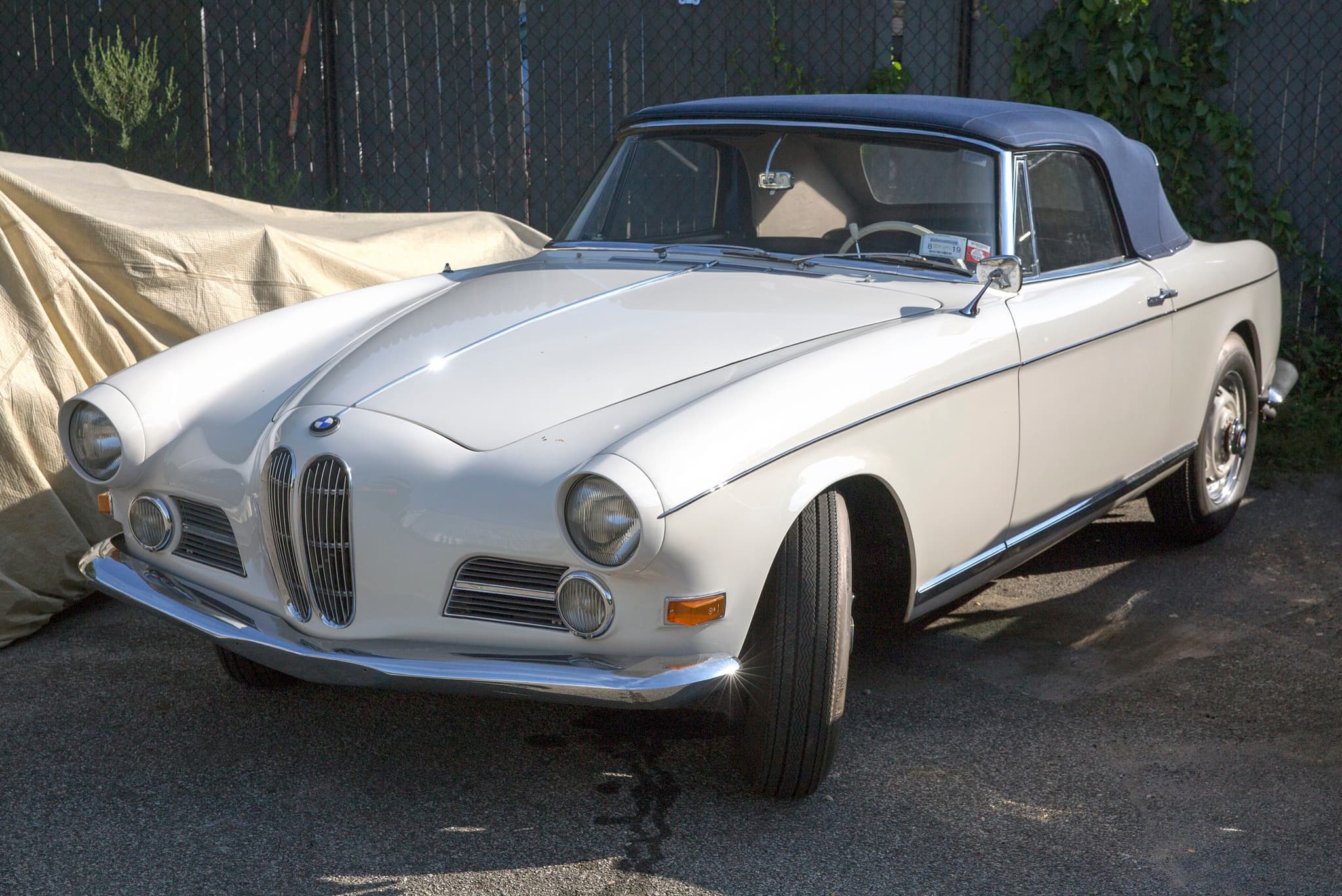 A white BMW 503 cabriolet with a blue fabric hood raised, parked on tarmac by a fence.