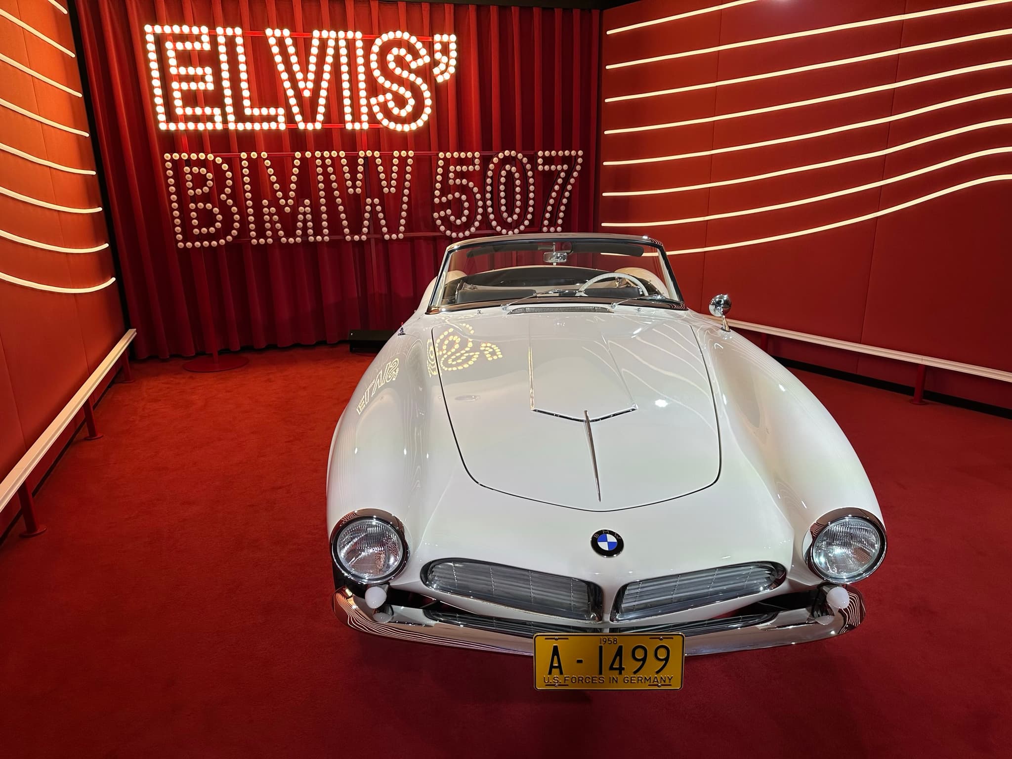 A white open two-seater seen head-on on a red carpet, its wide flat grille between two round headlamps and a yellow number plate below, with lit letters reading Elvis' BMW 507 on the red wall behind.