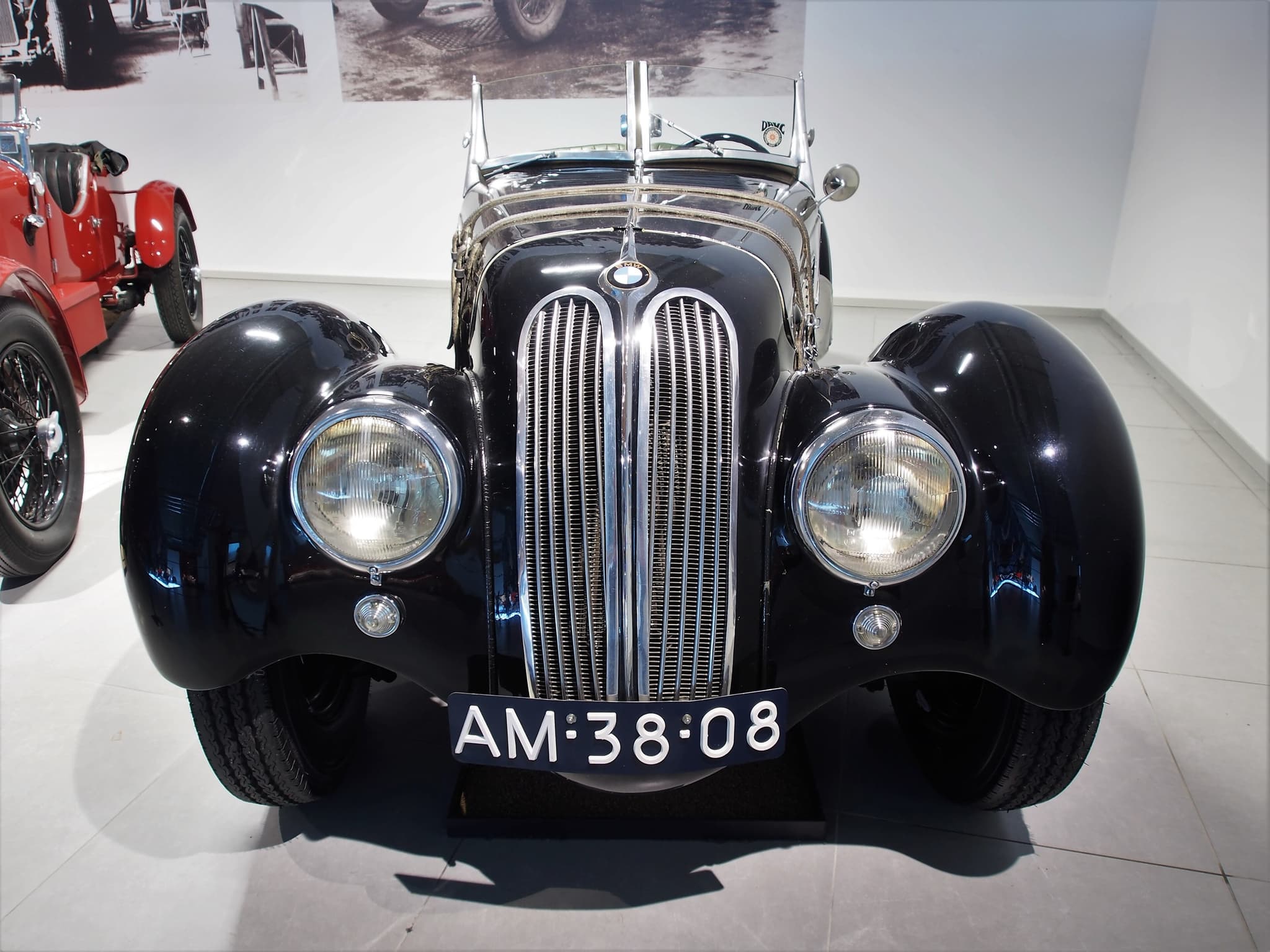 A black BMW 328 roadster seen head-on in a museum, with its narrow twin kidney grilles, headlamps set into the wings and a folding windscreen.