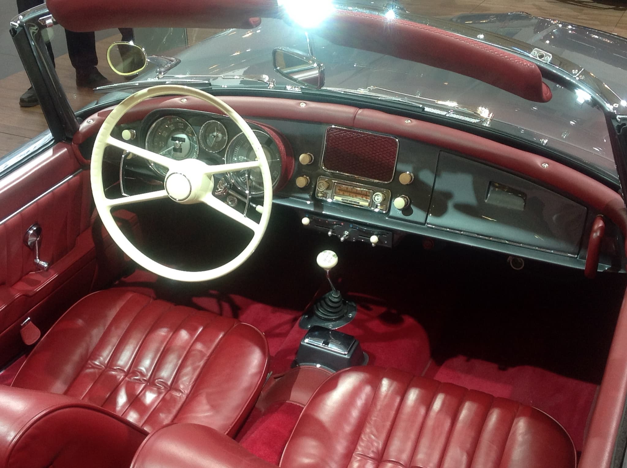 The cockpit of a grey BMW 507 with red pleated leather seats, an ivory steering wheel and a loudspeaker grille in the middle of the dashboard. Opens a larger view