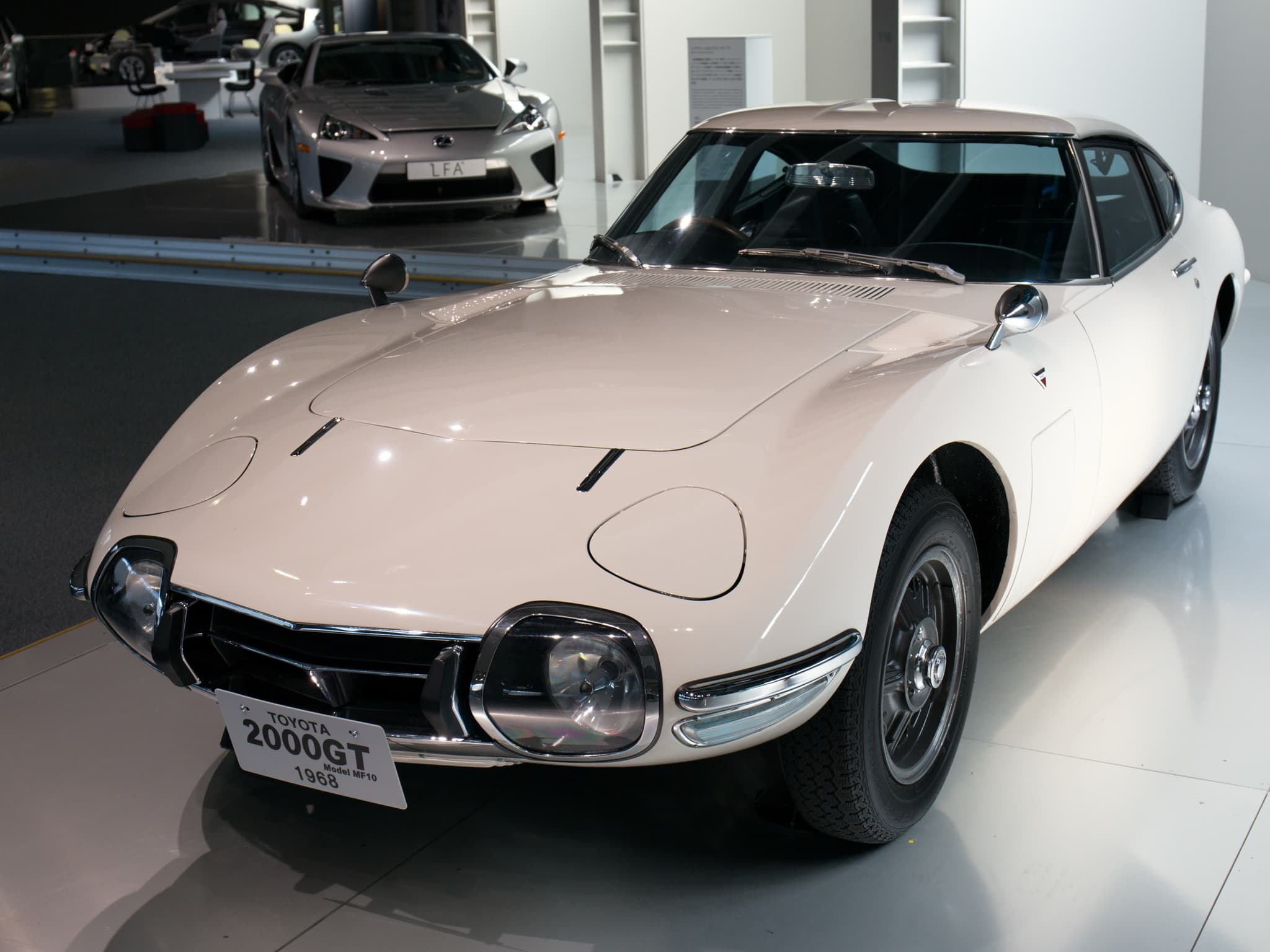 A white Toyota 2000GT coupé on a museum floor with a small placard on its front.