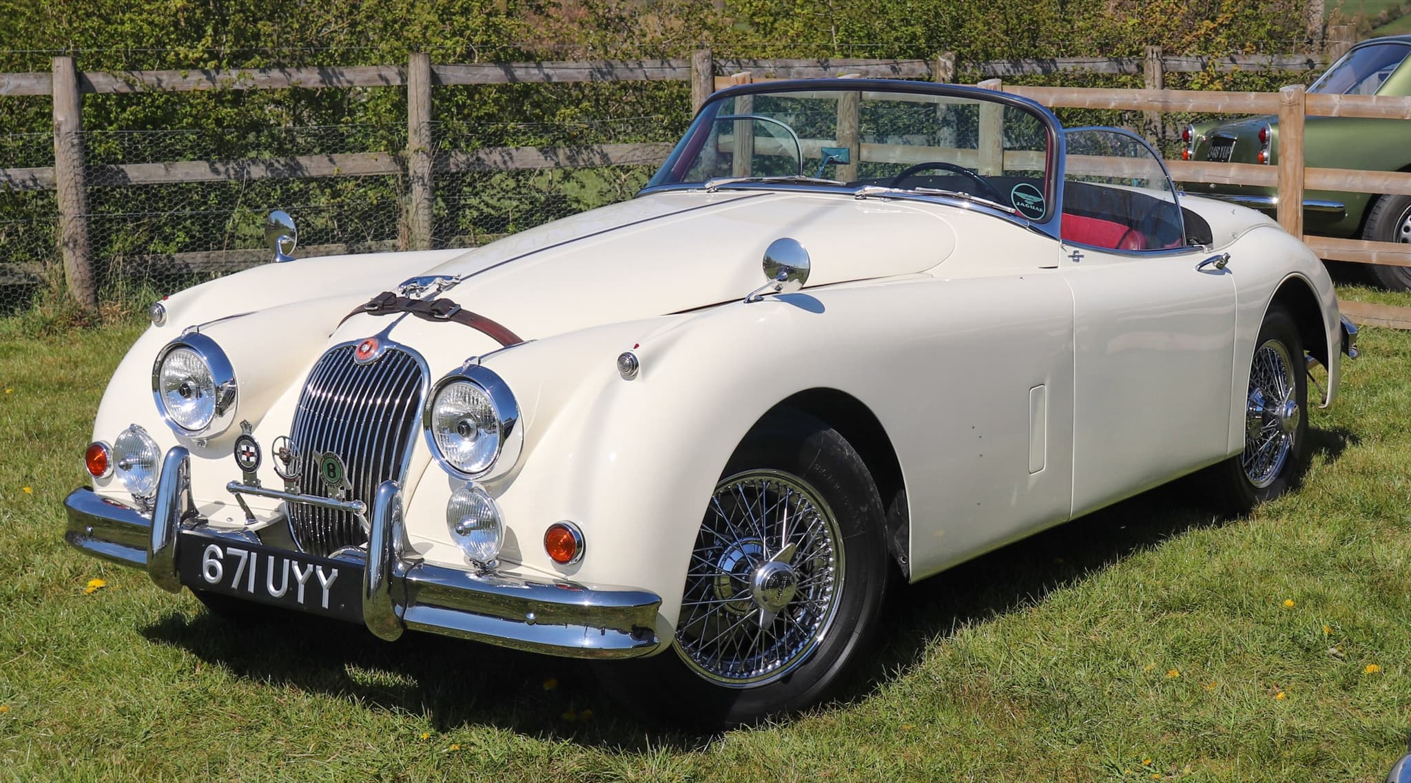 A white Jaguar XK150 roadster with a red interior and wire wheels on grass by a wooden fence.