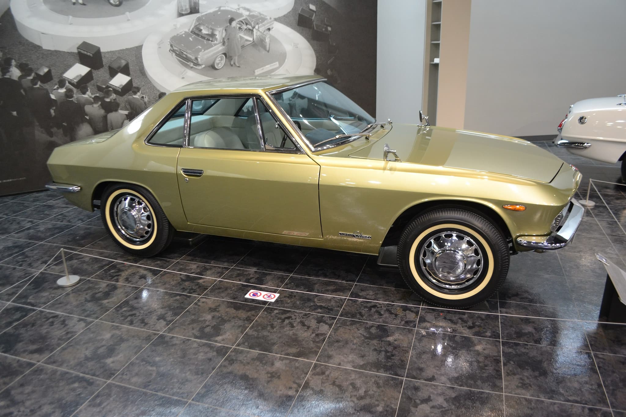 A gold Nissan Silvia coupé of the 1960s in profile on a dark tiled museum floor, with whitewall tyres.