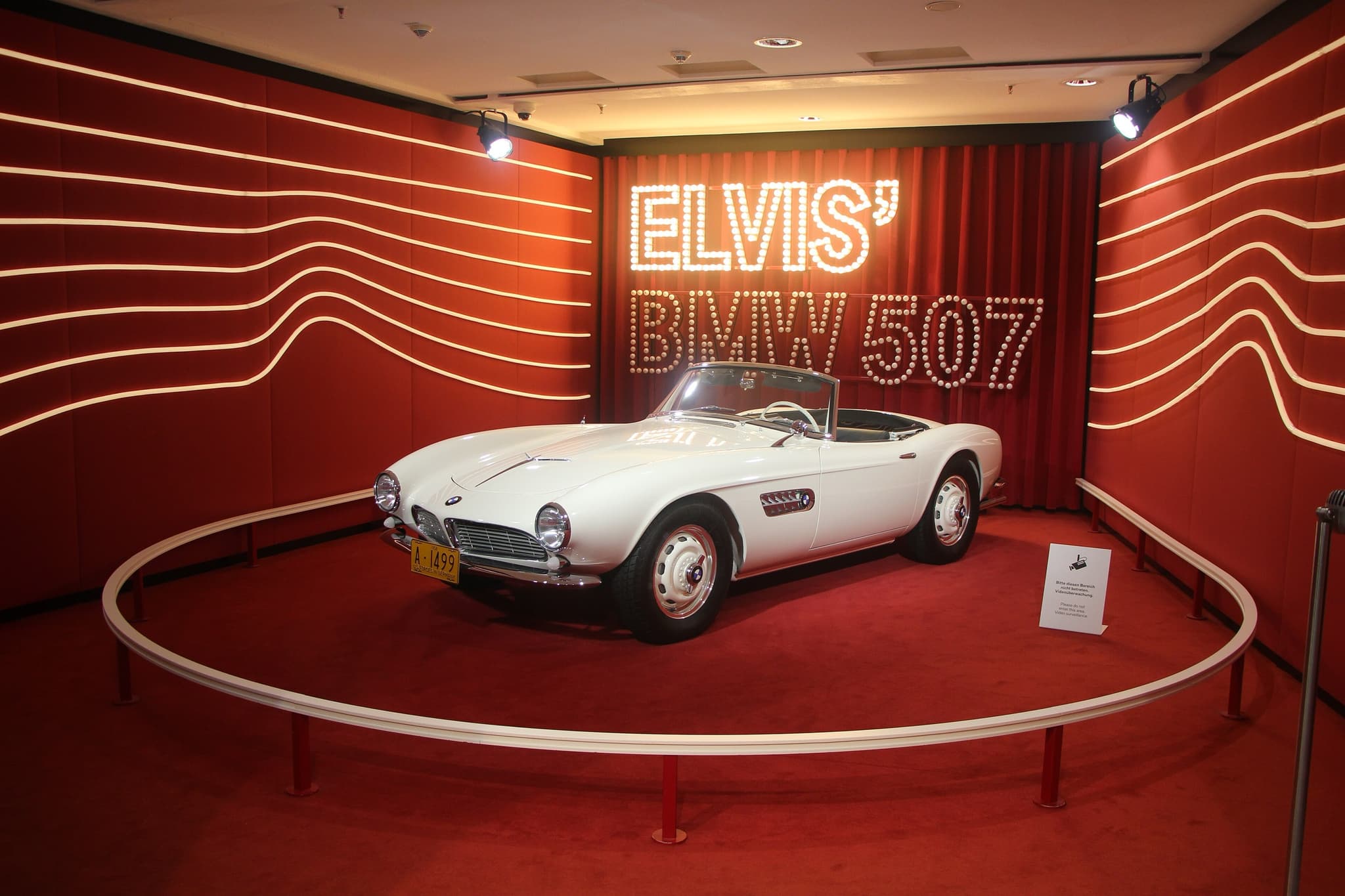 The white BMW 507 that belonged to Elvis Presley on a round red platform in a museum room, under a lit sign reading Elvis' BMW 507.