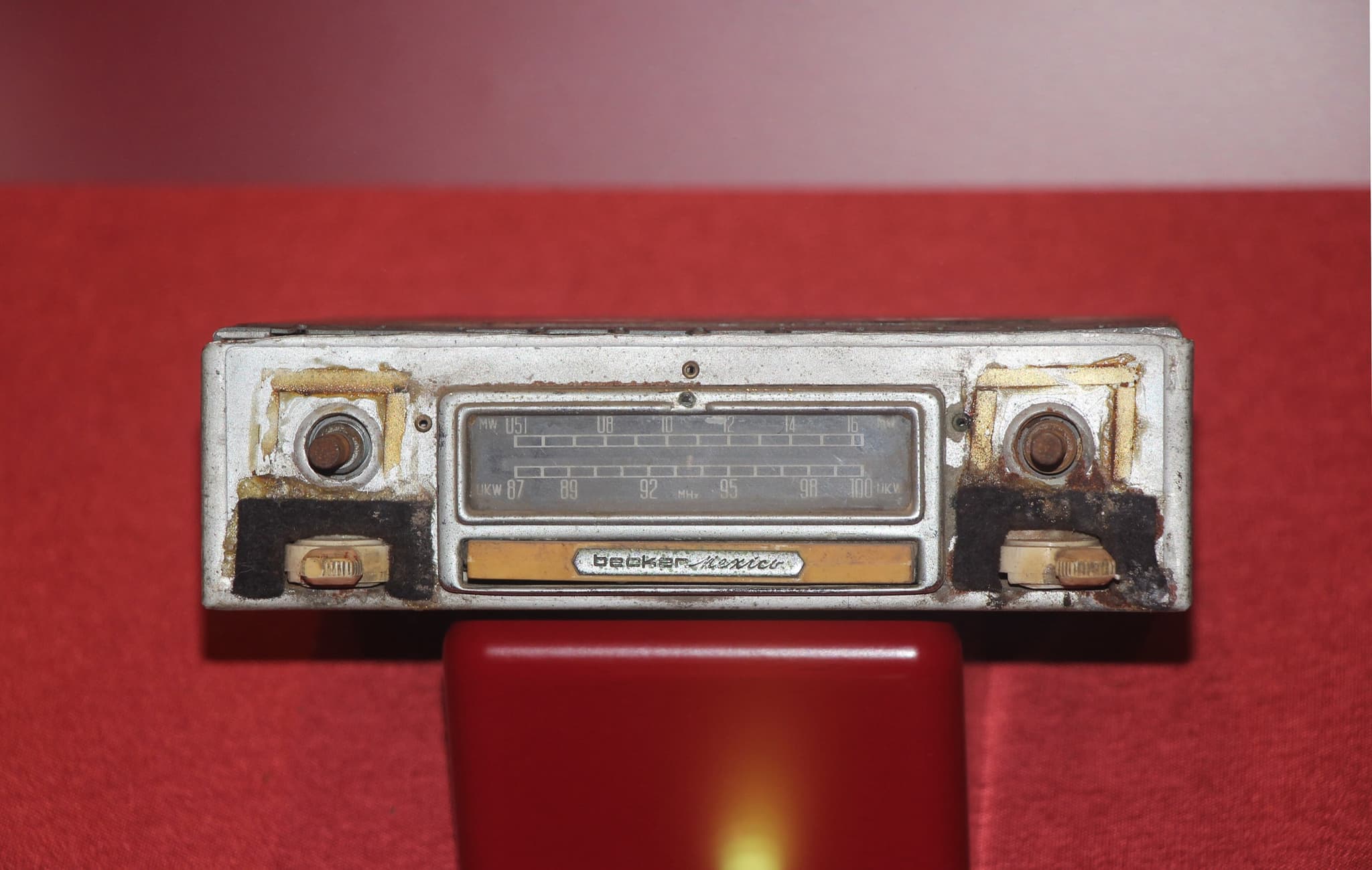 A worn chrome Becker Mexico car radio with a station dial and two knobs on a red display stand. Opens a larger view