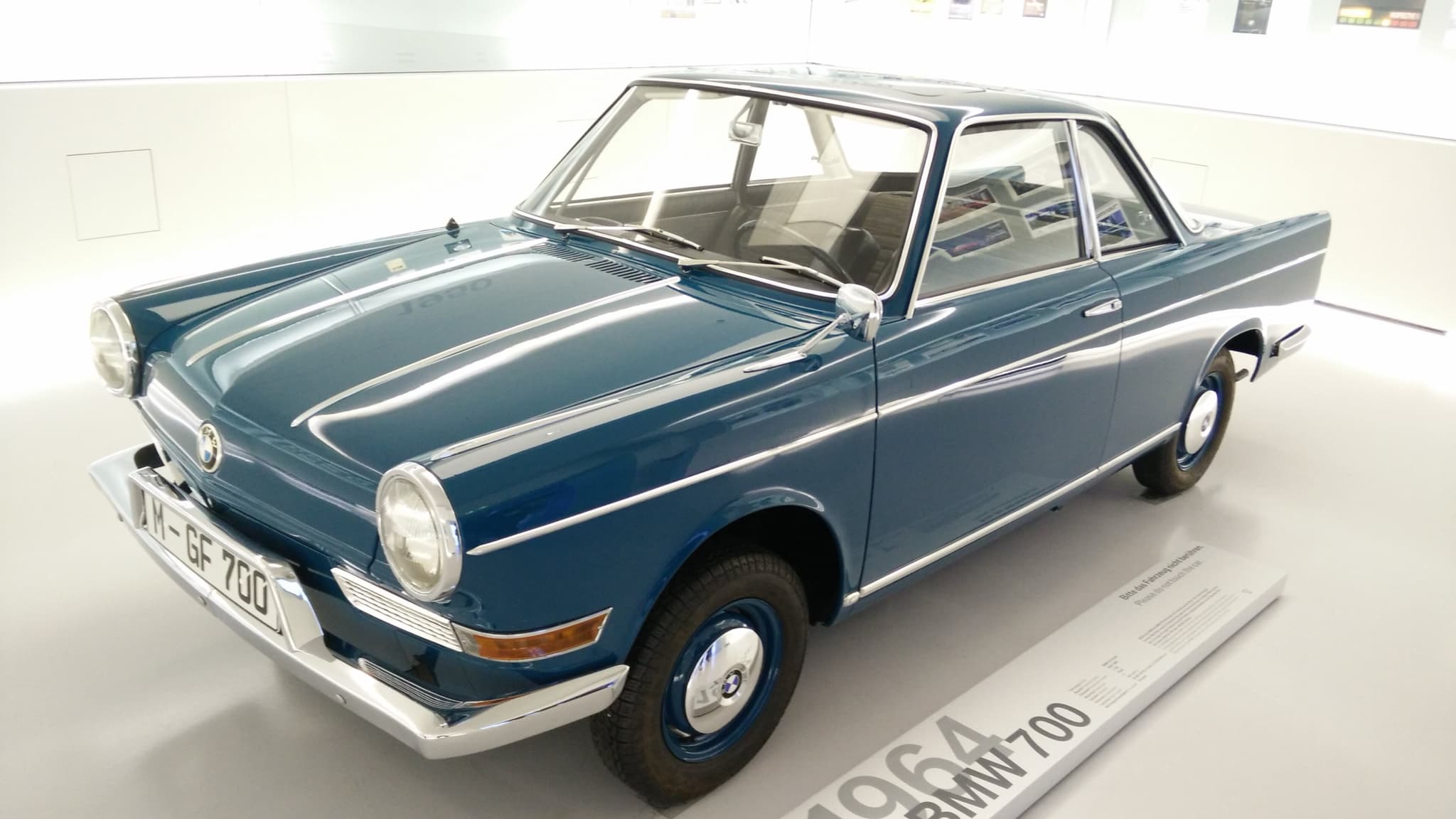 A blue BMW 700 LS coupé on a white museum floor beside a plinth reading 1964 BMW 700.