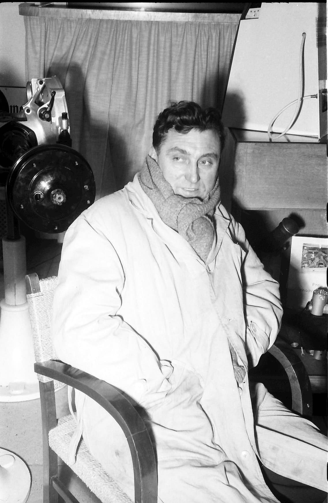 A black-and-white photograph of Ernst Loof in a raincoat and scarf, seated beside car parts.