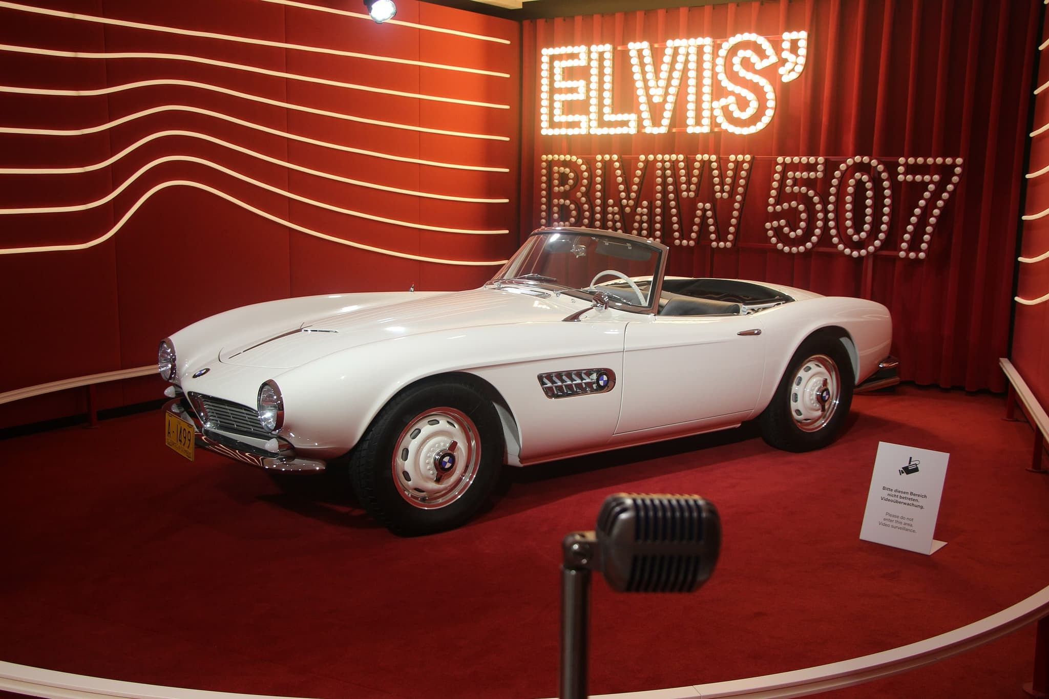 The white BMW 507 that belonged to Elvis Presley seen from the side on a red platform, with an old microphone on a stand in front.