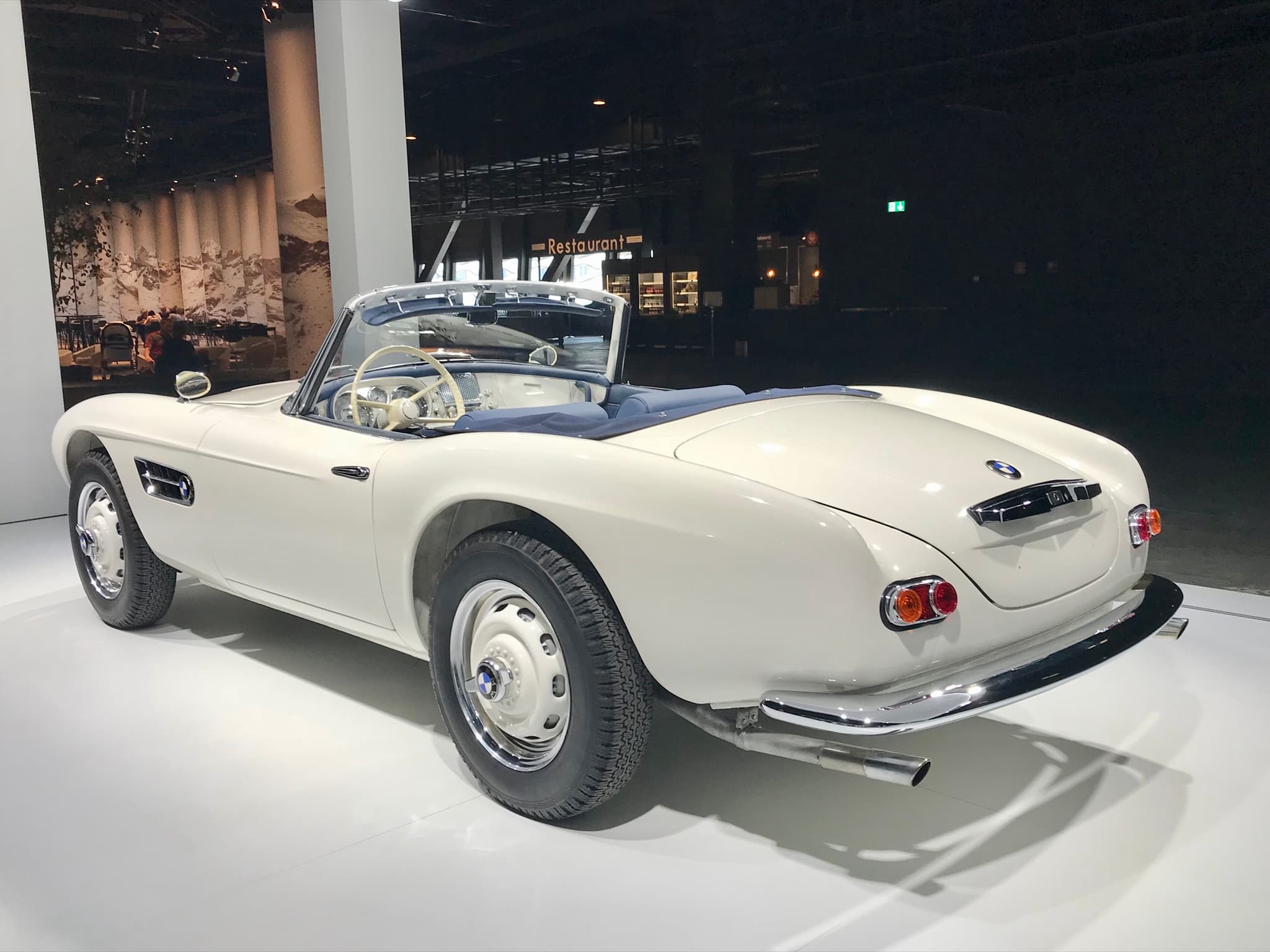 A white BMW 507 roadster seen from the rear left on a white show floor, its hood folded under a blue cover, with an ivory steering wheel and twin exhausts. Opens a larger view