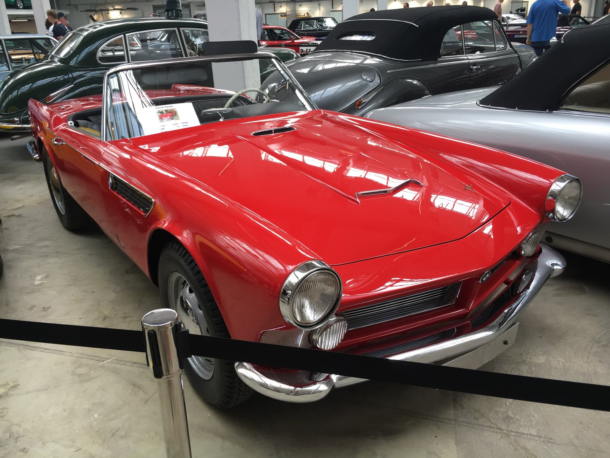 A red open two-seater with a low grille and round headlamps, the BMW 3200 Michelotti Vignale, behind a rope in a hall of classic BMWs with visitors behind it.