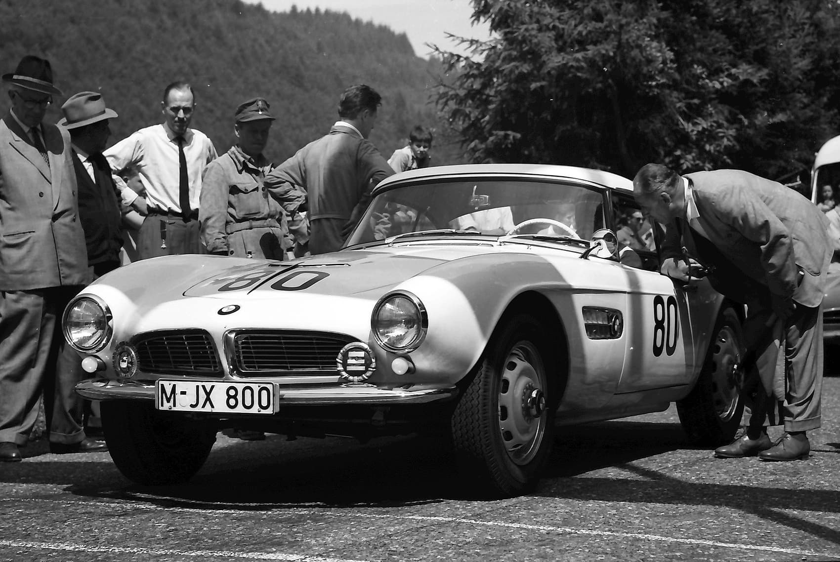 A black-and-white photograph of a BMW 507 with a hardtop and race number 80, registration M-JX 800, parked on a hill road while men in suits and caps stand around it.