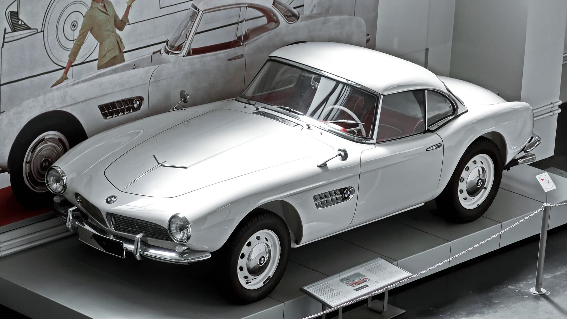 A white BMW 507 with a white hardtop on a museum plinth in front of a large wall drawing.