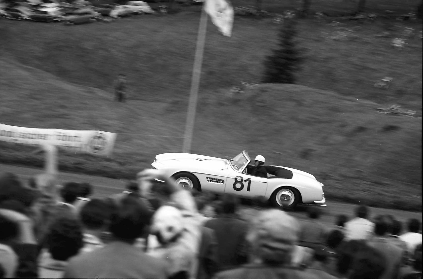 A black-and-white photograph of a BMW 507 roadster numbered 81 racing past a crowd of spectators on a hill climb.