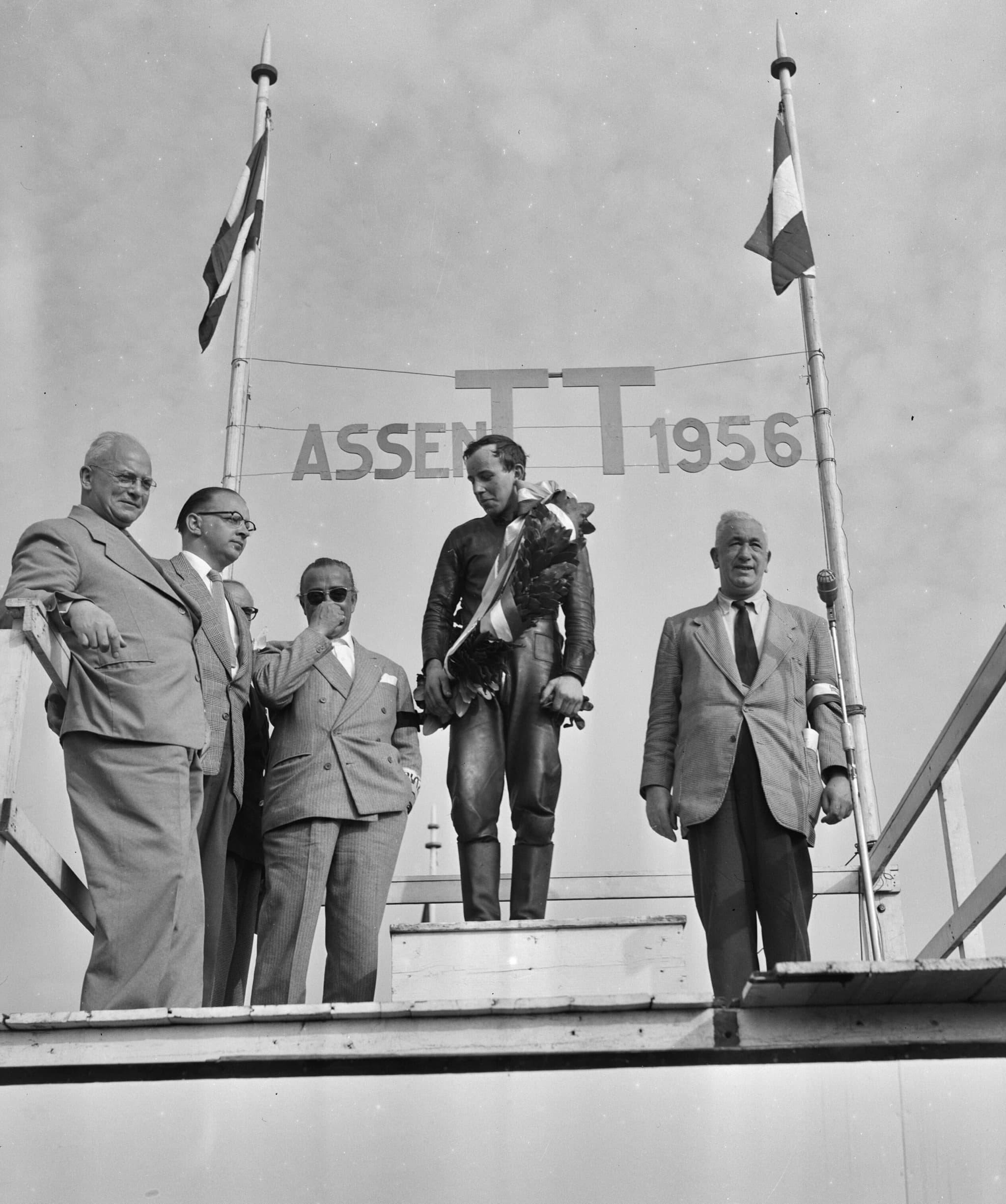 A black-and-white photograph of John Surtees in racing leathers holding a wreath on a podium under a sign reading Assen TT 1956, with five officials in suits.