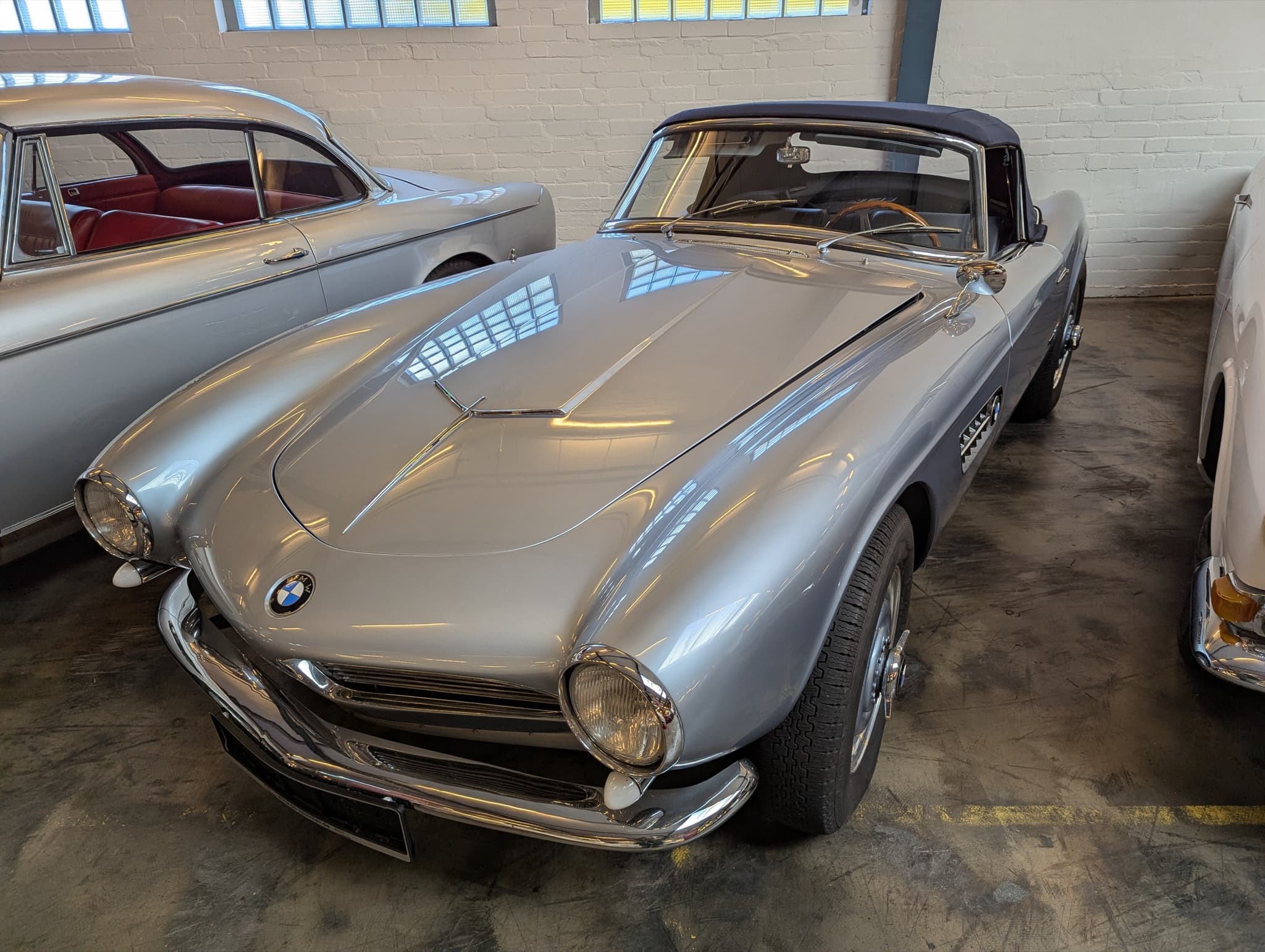 A silver BMW 507 roadster with a dark blue hood raised, in a brick museum hall.